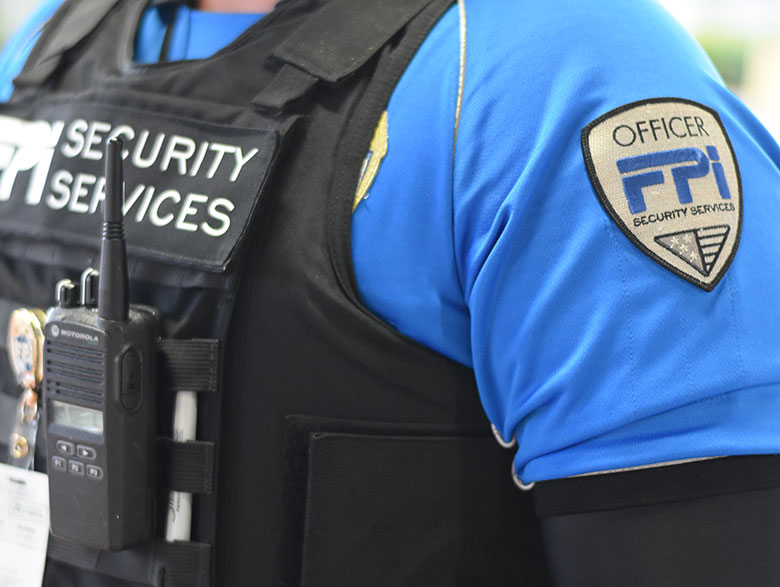 A close up picture of a uniformed FPI Security Services guard. The uniform is a light blue with black bullet proof best and the logo seen on the left shoulder.