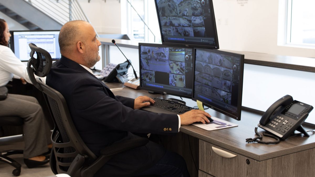 A member of FPI security services is sitting at a desk monitoring 3 screens that are recording different parts of the building. He is sitting in a black chair dressed in a black uniform on a sunny day.