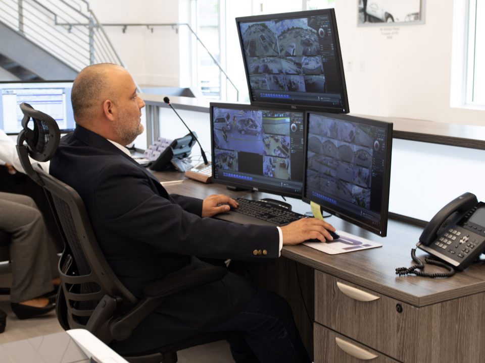 A member of FPI security services is sitting at a desk monitoring 3 screens that are recording different parts of the building. He is sitting in a black chair dressed in a black uniform on a sunny day.