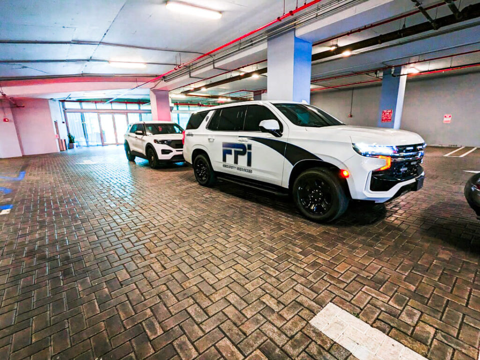 Two white vans in biscayne bay parking garage with the FPI Security Services logo on them. Both cars have their white headlights on.