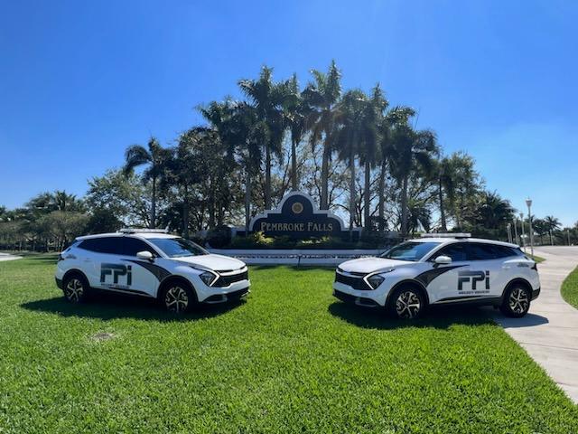 A sunny day with blue skies showing two white FPI Security Services vehicles parked on the green grass in front of the community sign of Pembroke Falls, located in Broward County.
