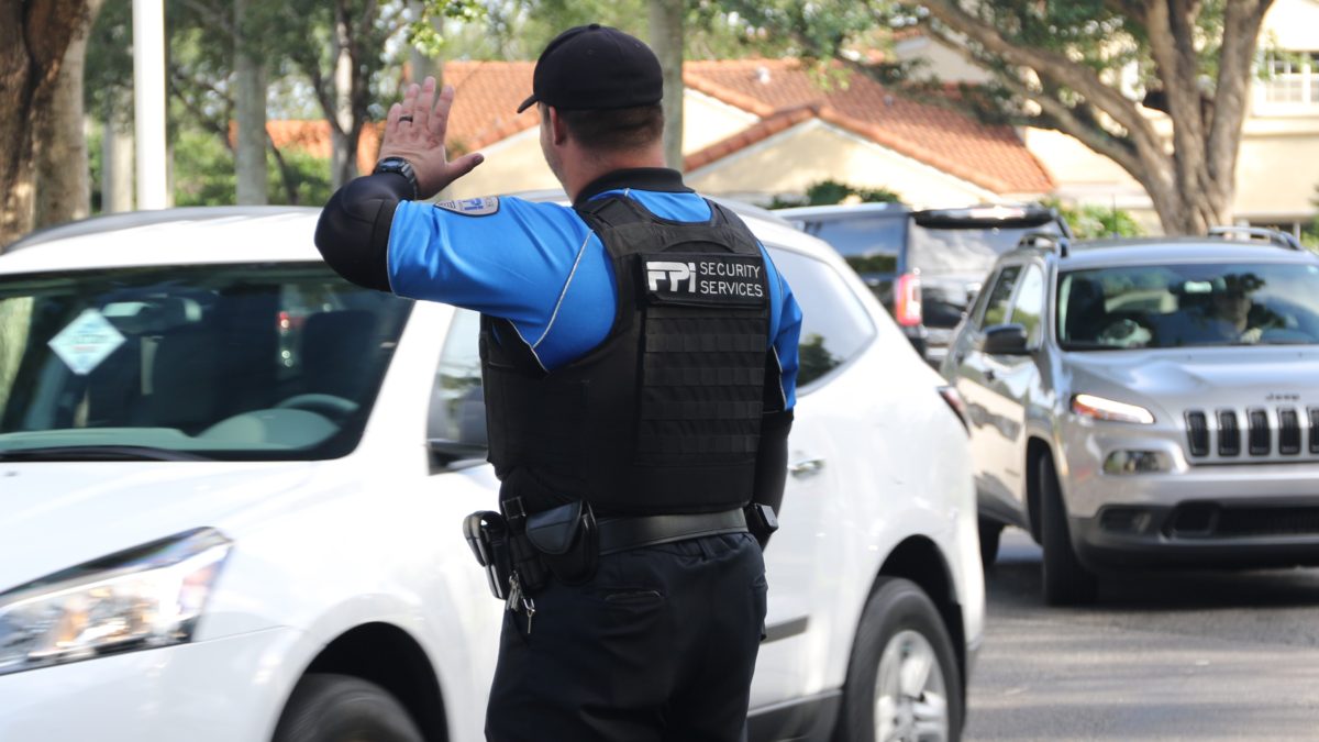 A black and blue uniformed FPI Security Officer standing outside helping traffic as cars pass by in the middle of a sunny day in the middle of a road.