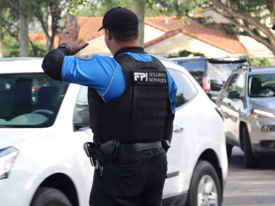 A black and blue uniformed FPI Security Officer standing outside helping traffic as cars pass by in the middle of a sunny day in the middle of a road.