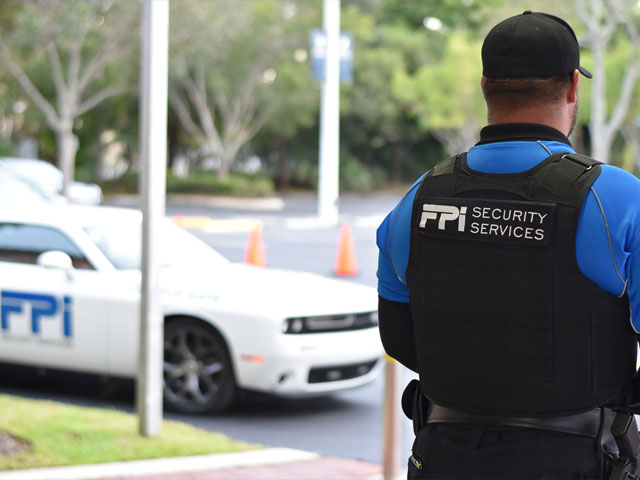 A white car with the FPI Security Services logo is parked on a grey road, next to some green grass in the middle of a sunny day. In front of the white car is a man in a blue and black FPI Security Services uniform facing the car and street.