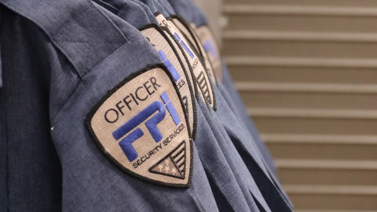 Blue shirts lined up on the left side of the image with the FPI security services logo on it. The logo is gold with the letters in blue and black. Behind the shirts are a set of stairs going up.