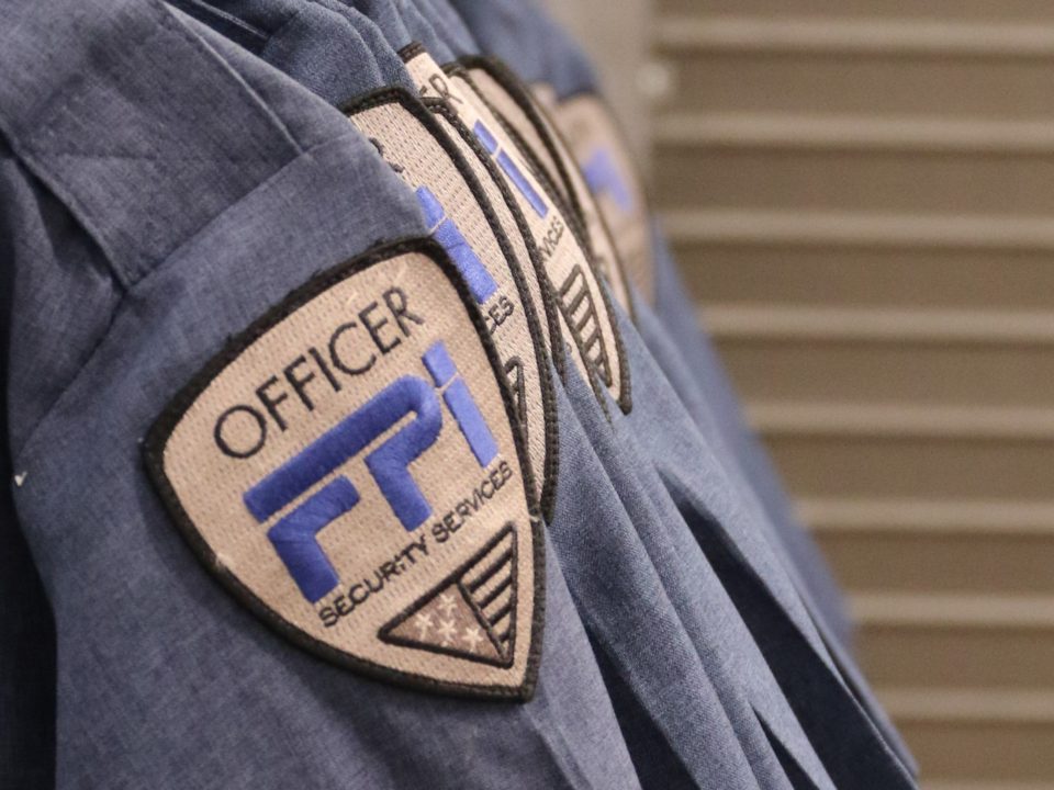 Blue shirts lined up on the left side of the image with the FPI security services logo on it. The logo is gold with the letters in blue and black. Behind the shirts are a set of stairs going up.