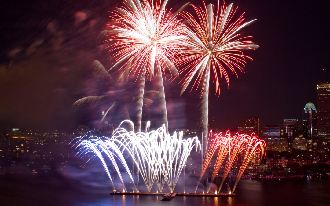 A picture of the night overlooking a body of water with red, blue and white fireworks going into the sky. The reflection can be seen in the water.
