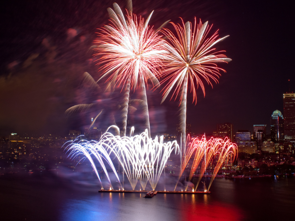 A picture of the night overlooking a body of water with red, blue and white fireworks going into the sky. The reflection can be seen in the water.