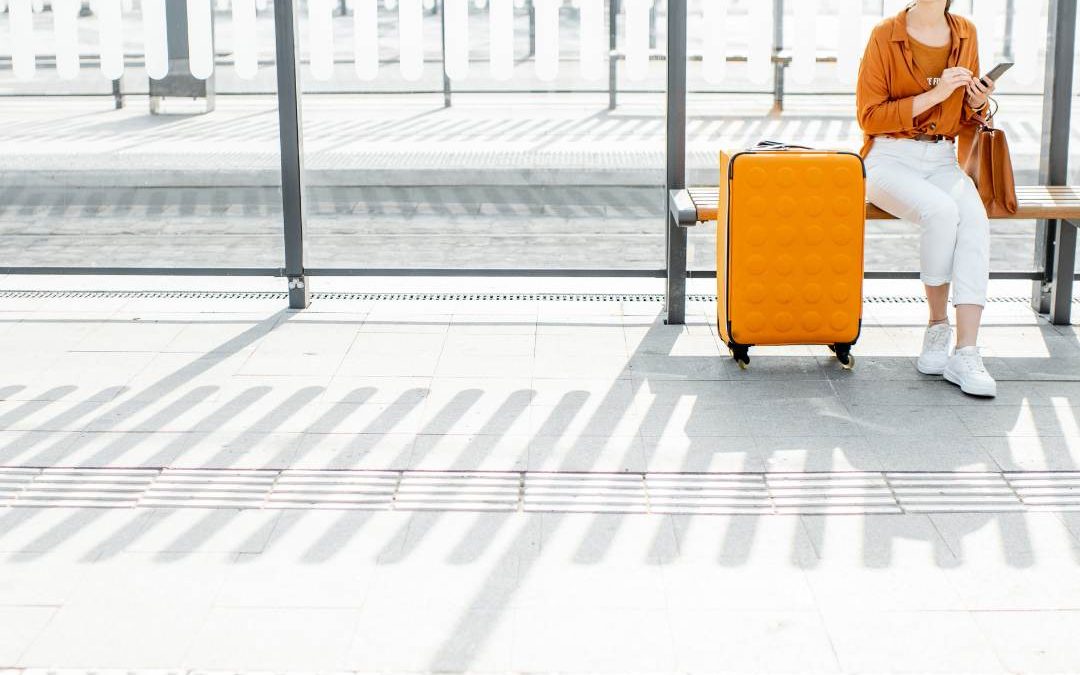 There is a lady in an orange and white outfit sitting on a white bench with an orange suitcase net to her, The background is a while bus stop area.