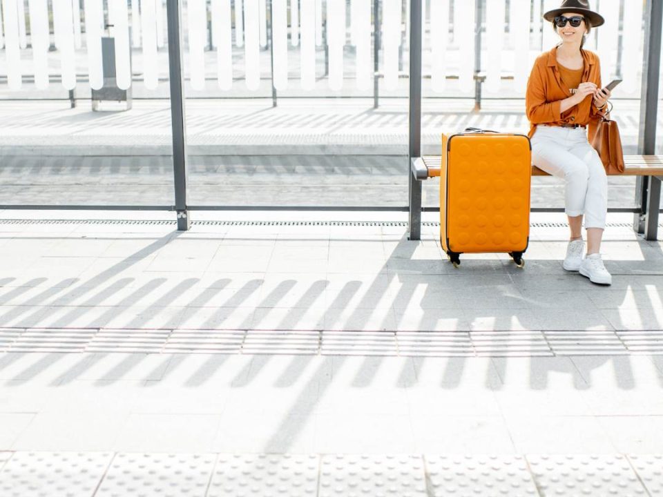 There is a lady in an orange and white outfit sitting on a white bench with an orange suitcase net to her, The background is a while bus stop area.