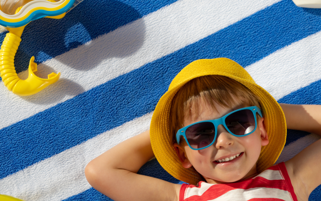 There is a child wearing a red and white striped shirt laying a blue and white towel with blue sunglasses on, They have a yellow bucket hag and have a snorkel mask next to them on this bright sunny day. This image is the cover for the following blog topic Summer and Back-to-School Tips for Child Safety.