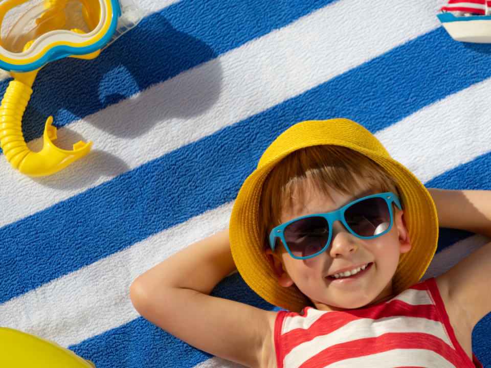 There is a child wearing a red and white striped shirt laying a blue and white towel with blue sunglasses on, They have a yellow bucket hag and have a snorkel mask next to them on this bright sunny day. This image is the cover for the following blog topic Summer and Back-to-School Tips for Child Safety.
