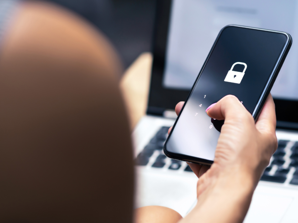 There is a picture of a lady holding a phone in her right hand, the cell phone is black with a white lock on it. in the background there is a laptop