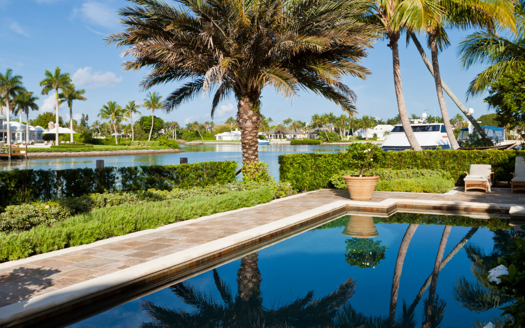 There is an outdoor picture of ta back of a house with a pool overlooking a body of water. It's a sunny day with a boat on the water and some palm trees along the edge of the blue pool.