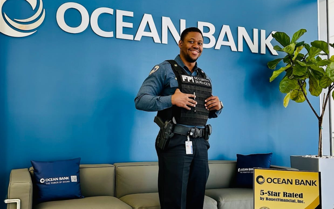 This is a picture of and armed FPI Security Services guard standing in front of an ocean bank sign. The wall is blue and there is a couch in front of it.
