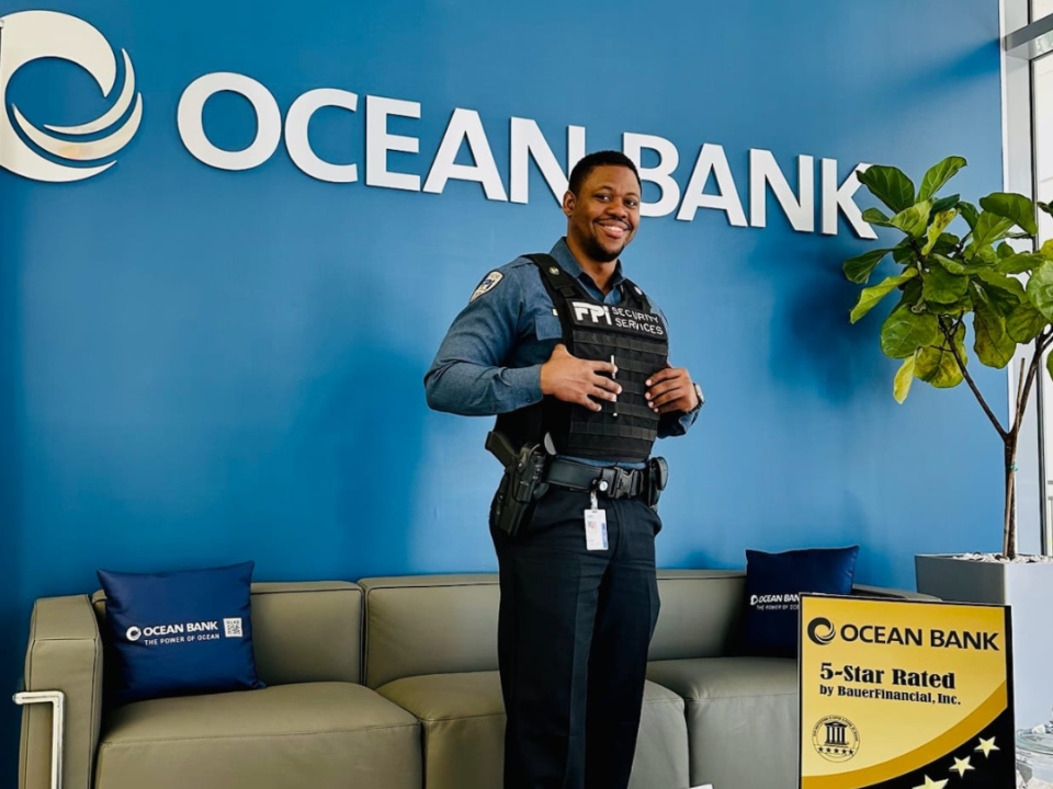 This is a picture of and armed FPI Security Services guard standing in front of an ocean bank sign. The wall is blue and there is a couch in front of it.