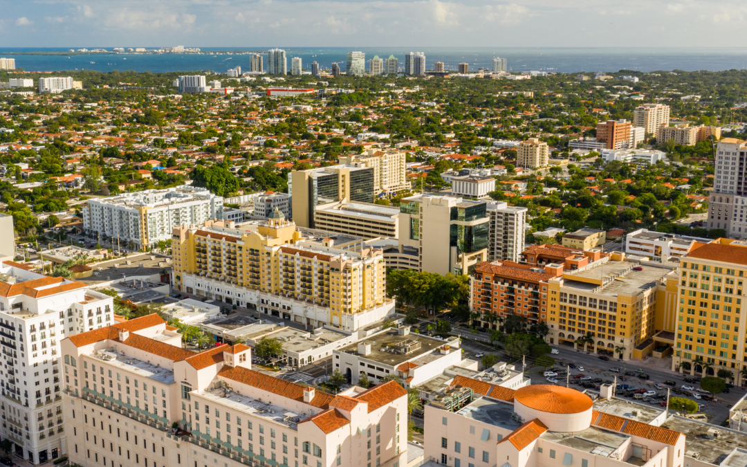 This is a picture of an overview of the buildings in coral gables florida, There are white and yellow buildings and there are some trees in the background as well. The sky is blue.