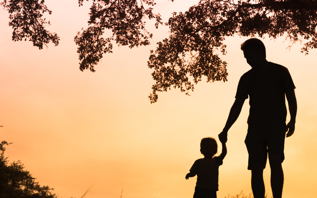 This is an image with a yellow background as the sun sets with a dark silhouette of a child holding the hand of an adult man as they walk under some trees.