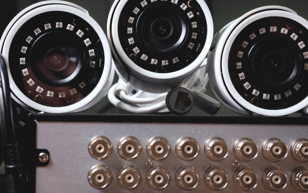 Image shows three security cameras that are white stacked next to eachother. There is a dark brown background on top of a console with several inputs for chords.