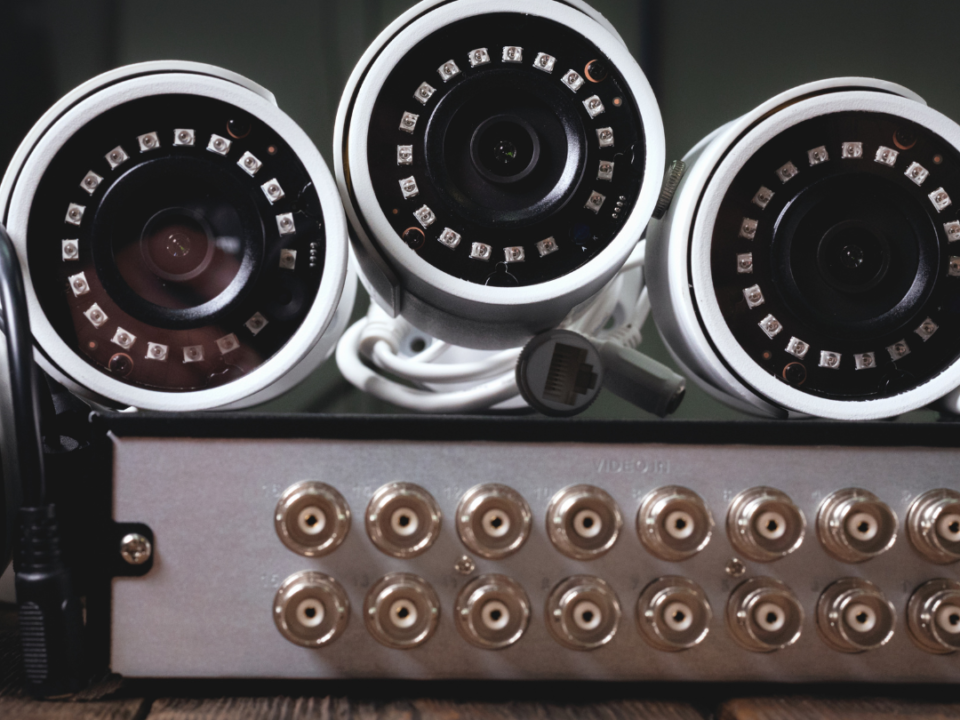 Image shows three security cameras that are white stacked next to eachother. There is a dark brown background on top of a console with several inputs for chords.