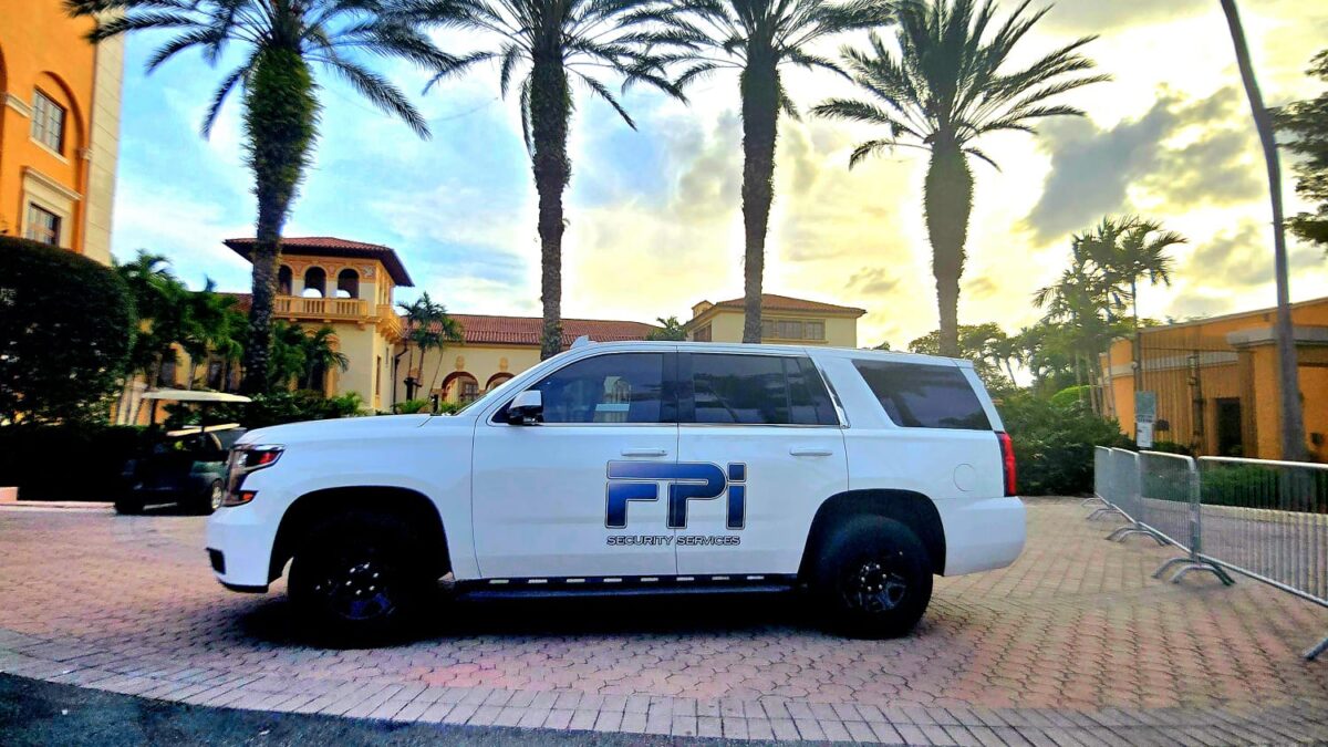 This is an image of a white SUV with the words FPI Security Services across the vehicle. It is shown parked to the side of the Biltmore Hotel, in Coral Gables. Behind the white car there are 5 palm trees as the sun sets showing hues of blue and yellow.