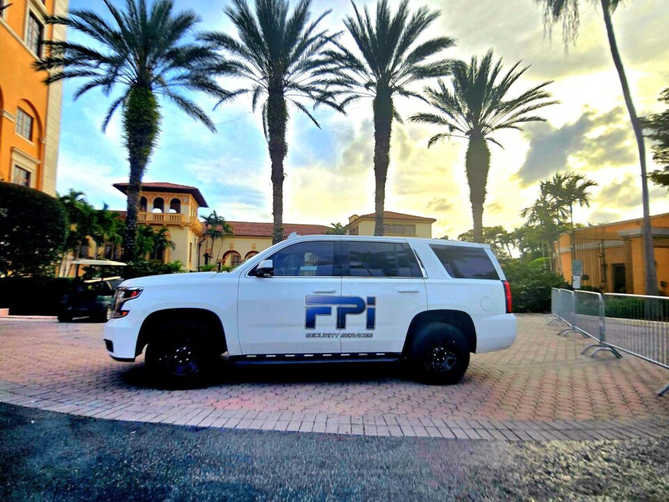 This is an image of a white SUV with the words FPI Security Services across the vehicle. It is shown parked to the side of the Biltmore Hotel, in Coral Gables. Behind the white car there are 5 palm trees as the sun sets showing hues of blue and yellow.