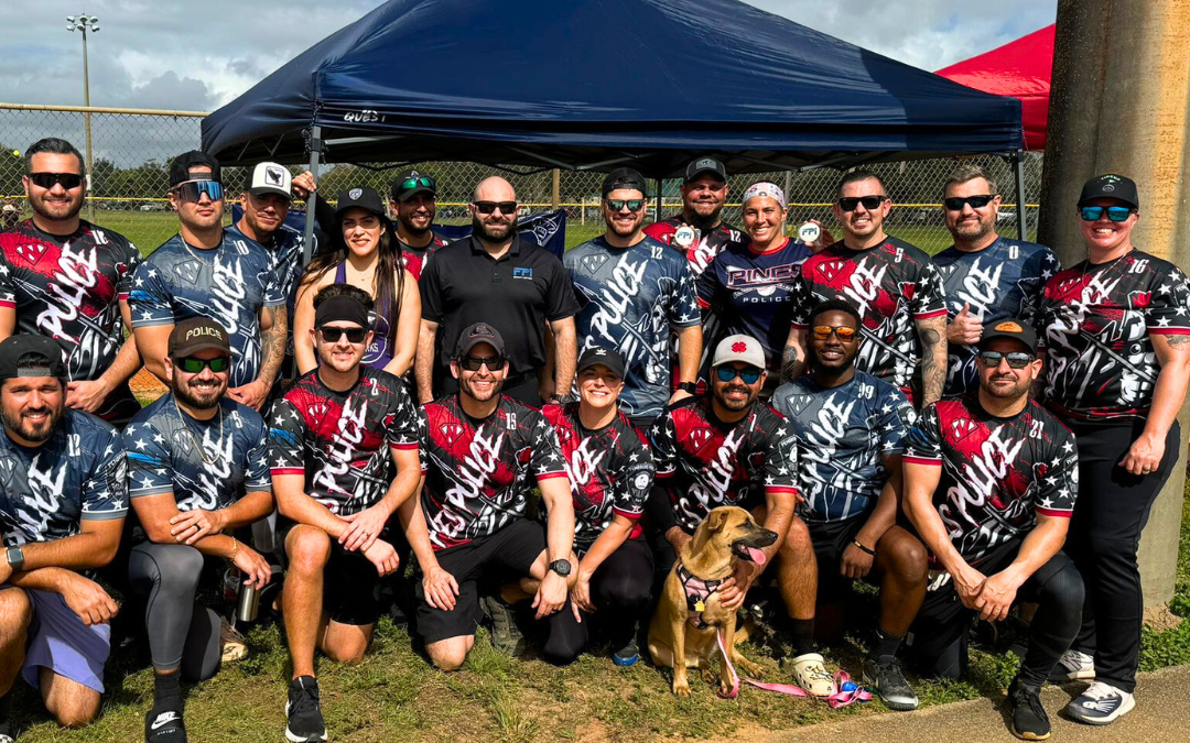 This is a group photo of the pembroke pines police department outside of a softball field. They are wearing their uniforms which are red white and blue and sitting in a group behind the FPI security services tent. In the picture there is the VP of the company and also the marketing manager.