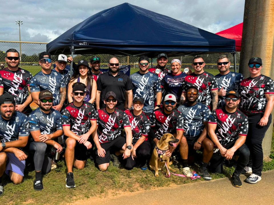 This is a group photo of the pembroke pines police department outside of a softball field. They are wearing their uniforms which are red white and blue and sitting in a group behind the FPI security services tent. In the picture there is the VP of the company and also the marketing manager.