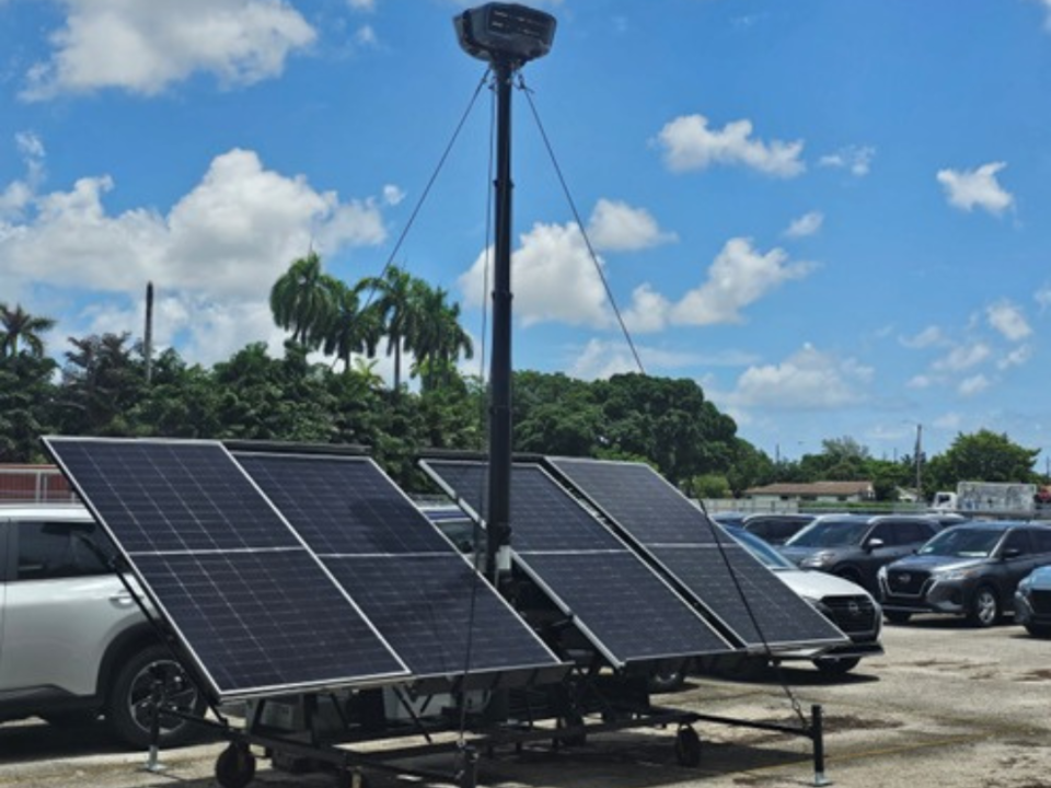 This is an image of the RIO360 security solution with solar panels and two cameras on top of a tall pole in the middle of a car lot. With a sunny day in the background and blue skies.