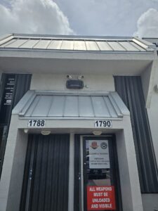 Florida Gun Center entrance with a ROSA attached to it. The building is grey with black doors. The background is of a light blue as well. 