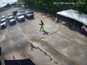 Image of a man walking with a red skateboard from a RIO360 camera in sunlight. The parking lot shows some silver cars parked to the top left of the image as well. 